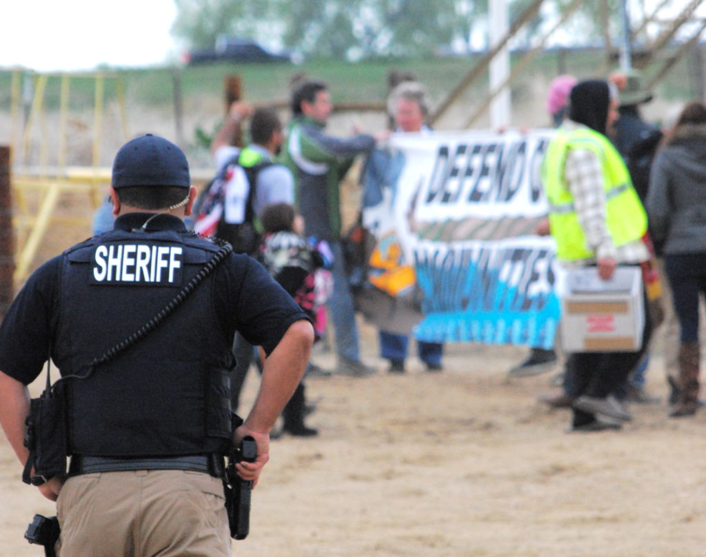 This photo was taken by Sam Dyer: Once marchers left the pavement and started down the private dirt road towards the well site, they were told they would be arrested. They were not. After ignoring commands to stop and continuing past police to the well, the officers drew a new line in the sand. Protesters were told they would definitely be arrested if they climbed onto the spill-protection dirt embankment surrounding the tanks. 