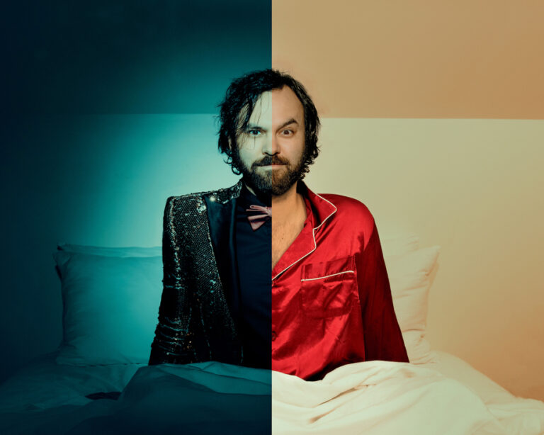 A bearded man, the musician known as Shakey Graves, sits up in bed — half his body is dressed in pajamas, the other half in a suit.