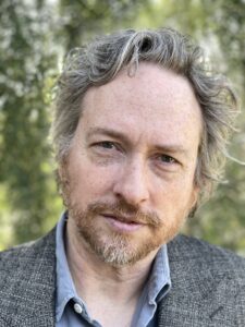 The headshot of a man, screenwriter Zeb Wells, with gray hair and a goatee. 