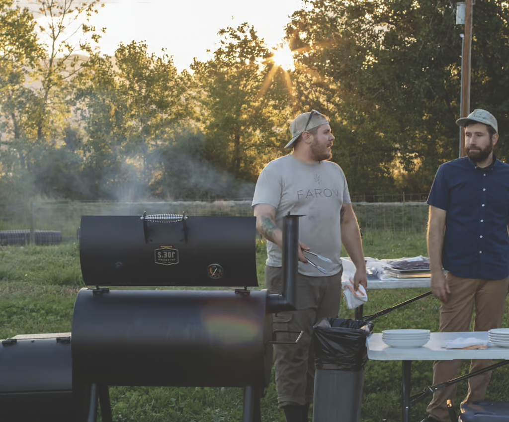 two men standing at a smoker grill outside on grass and trees at sunset