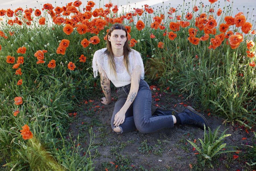 A woman with long brown hair, a white shirt and jeans sits in the dirt among a small field of roses.