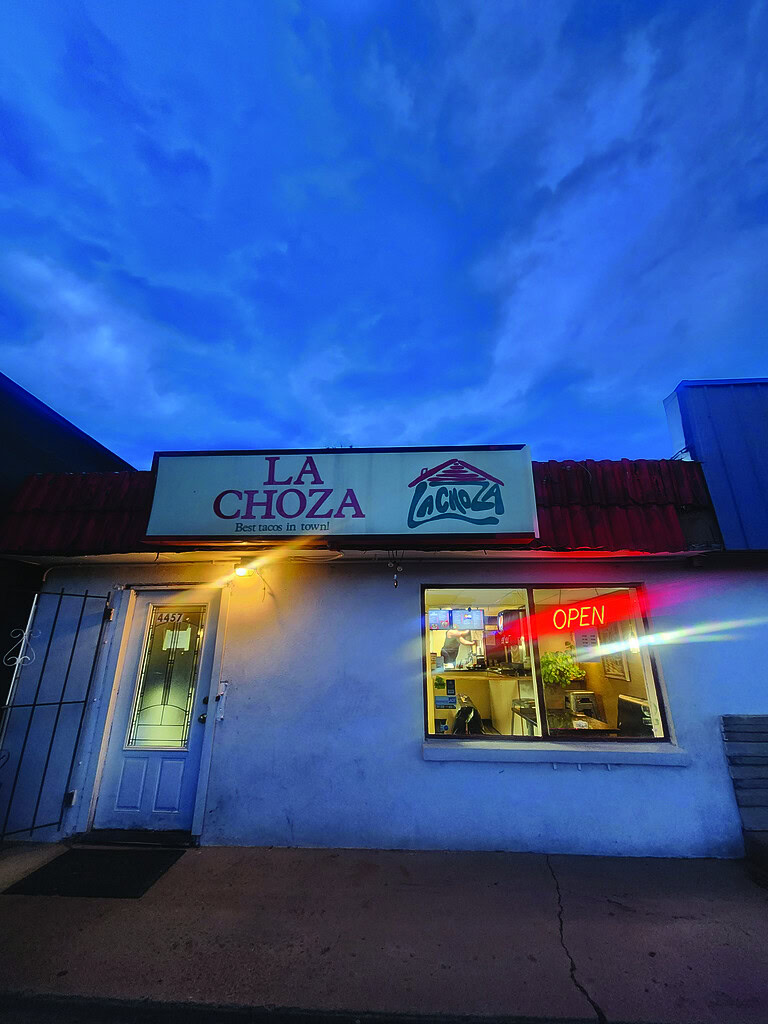 dingy white building with red roof against dark blue cloudy ski with neon red open sign in the window and La Choza name