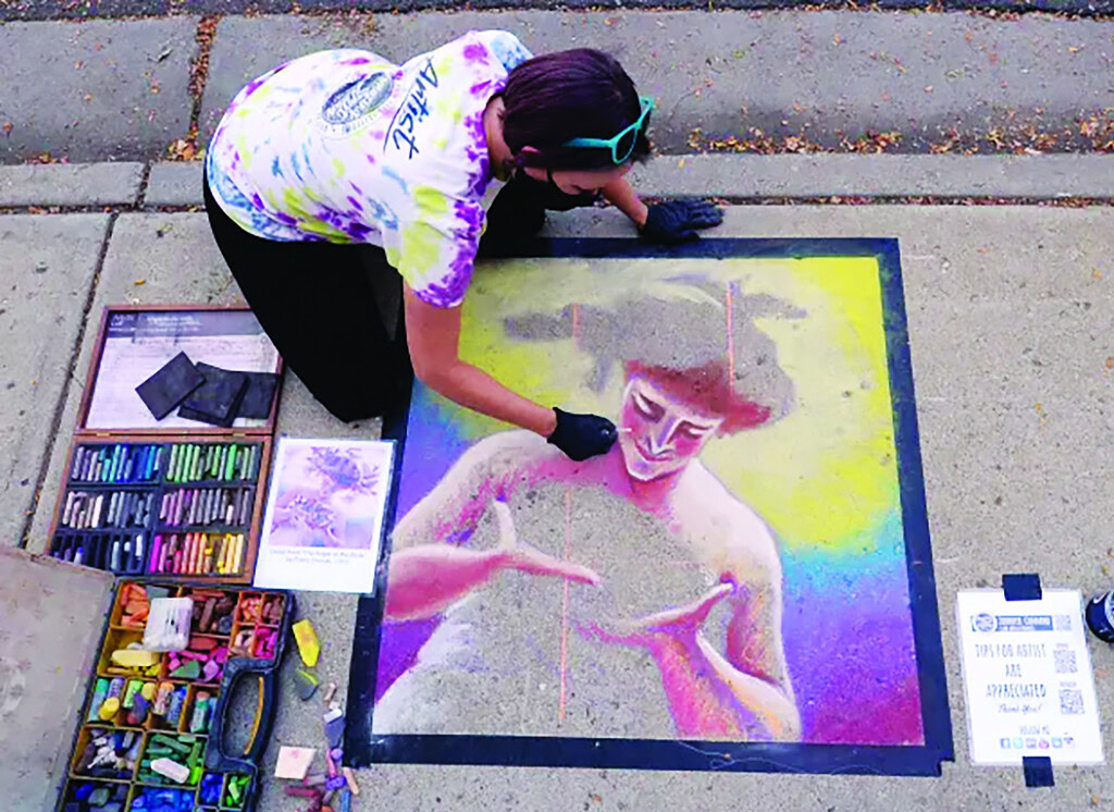 person in tie dye shirt drawing multicolored chalk mural on sidewalk