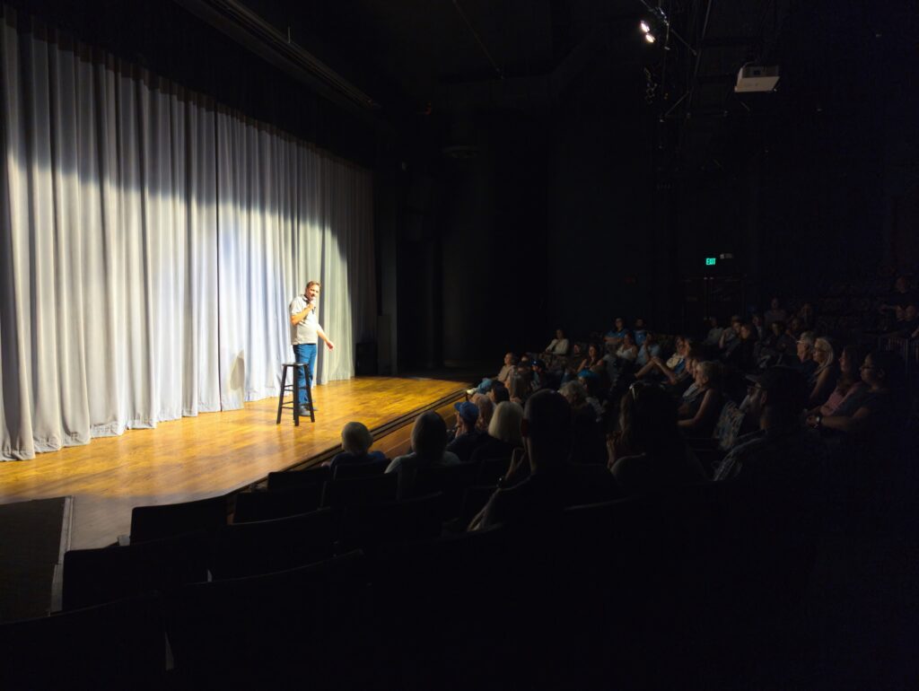 man comedian on stage with light curtains wood floor and audience in shadow